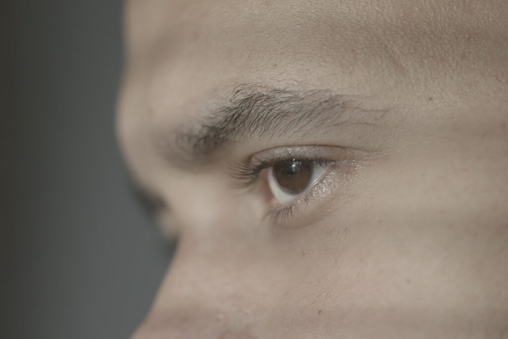 Bilden föreställer en närbild på en persons öga, ögonbryn och näsrot i profil som delvis skuggas av fönsterpersienner. 